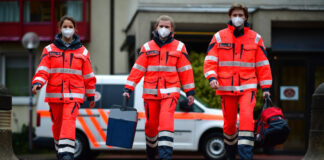 Drei gehende Johanniter Mitarbeiter mit Maske vor Einsatzwagen