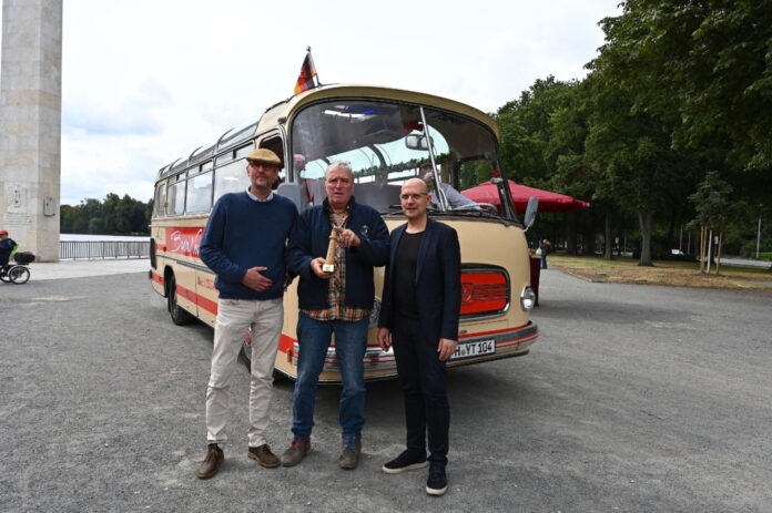 Die BussaNova Classic Tours-Inhaber Thorsten Stahl (v. l.) und Torsten Krüger nehmen von Jens Palandt, Klimadezernent der Region Hannover, den symbolischen Leuchtturm für die Förderung des Bus-Umbaus entgegen.