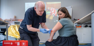 Ein Erste Hilfe-Trainer zeigt am Beispiel einer Teilnehmerin in einem Erste Hilfe-Kurs, wie man einen Verband am Ellenbogen anlegt.