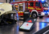 neue notfallsoftware soll feuerwehrkräften hilfe sichern feuerwehrhelm, funkgerät und smartphone als symbole für die notfallalarmierung. ki-generiertes symbolbild mit fiktiver bildschirmansicht.