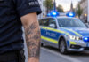 ki-generiertes symbolbild im querformat: tätowierter arm, kein gesicht, streifenwagen mit blaulicht im hintergrund.